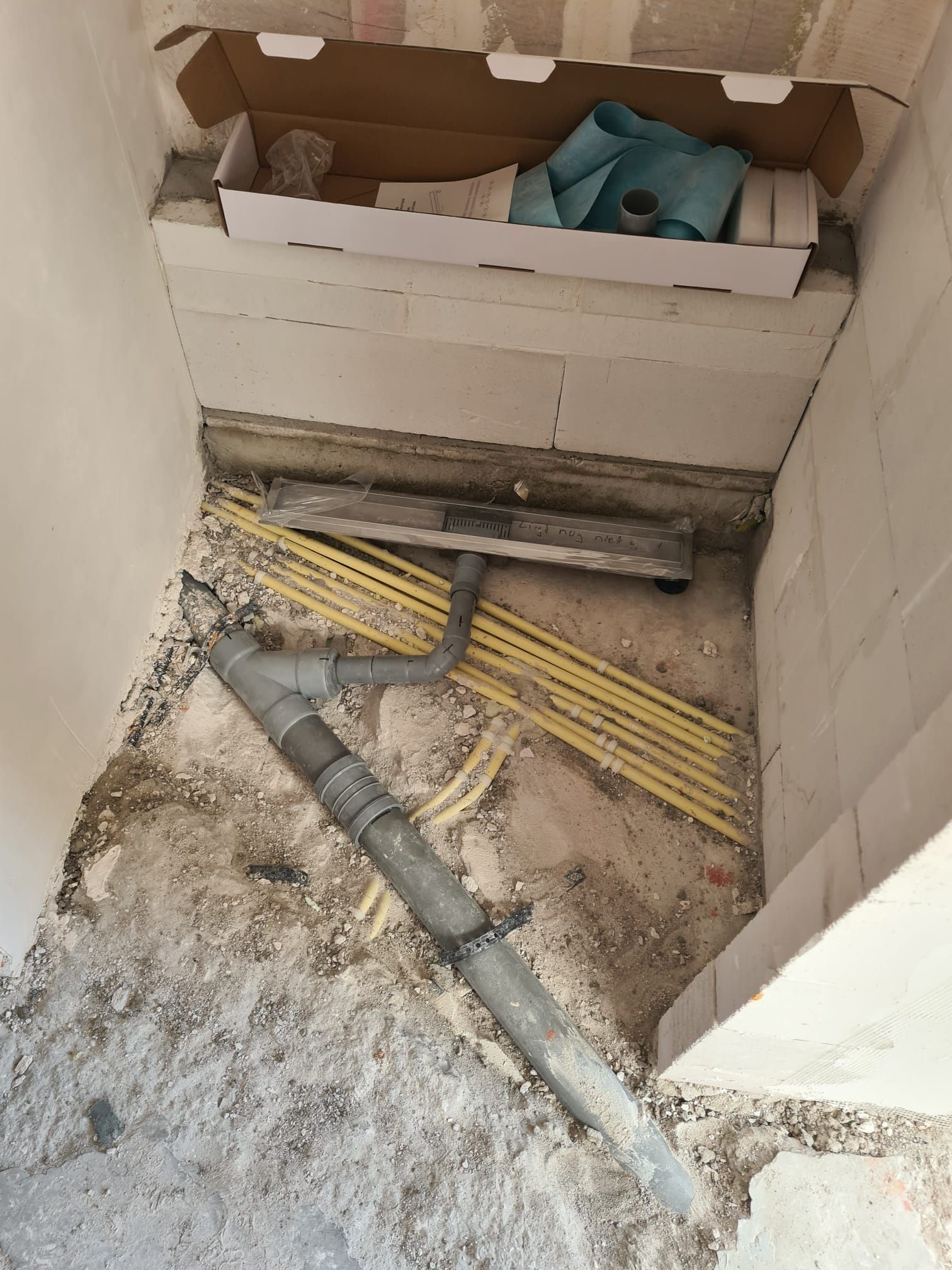 Een onafgewerkte bouwplaats met een grijs leidingsysteem op een betonnen vloer en een kartonnen doos met blauwe materialen.