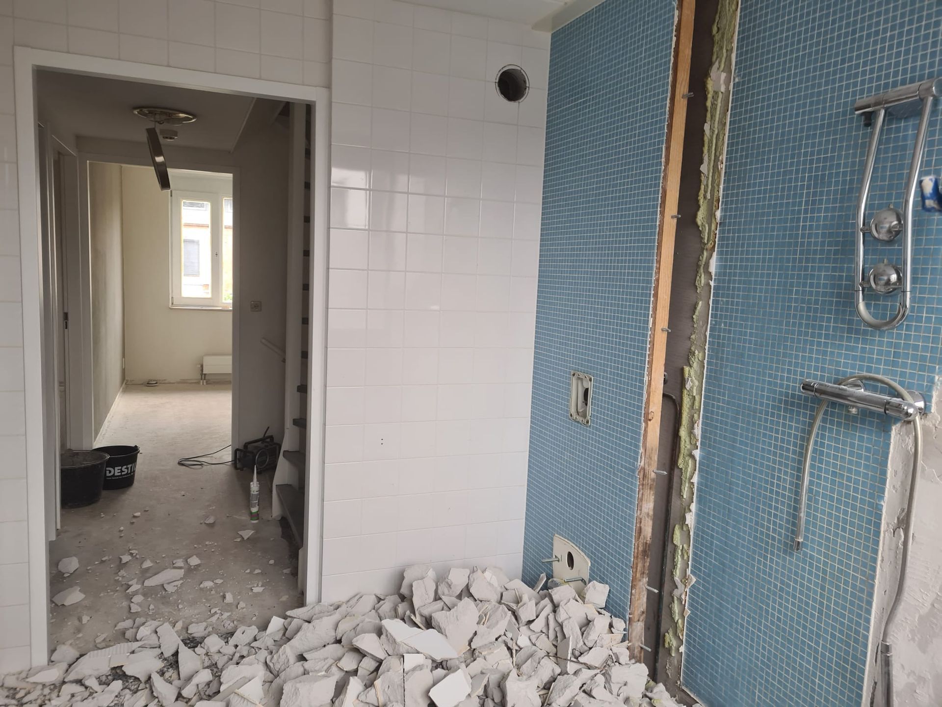 Een kamer die gerenoveerd wordt, met witte tegelwanden, een gedeelte met blauwe tegels met een patroon en bouwafval op de vloer.