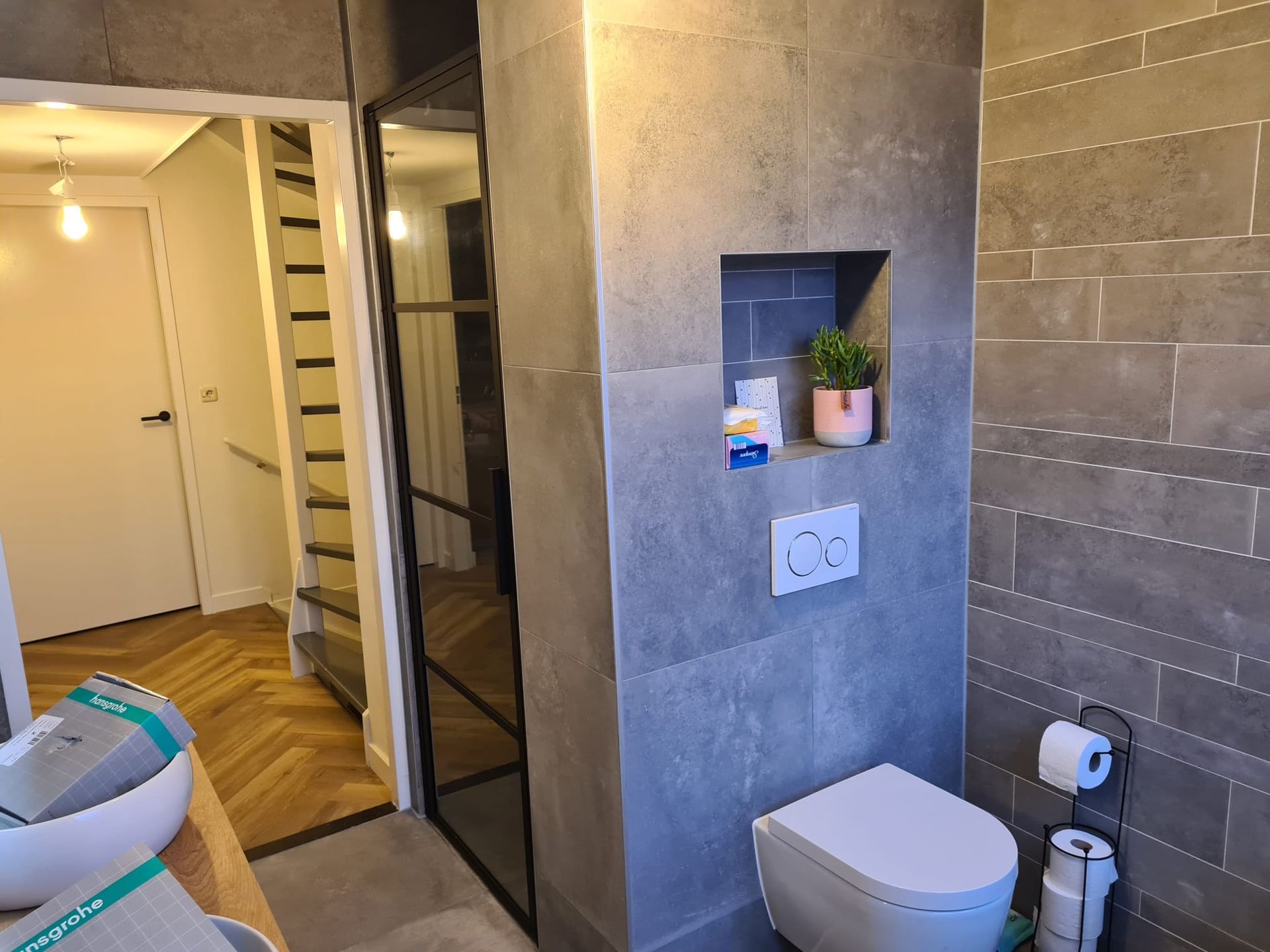 Een wandtoilet en een ingebouwde plank in een badkamer met grijze tegelwanden, grenzend aan een deuropening en een trap.