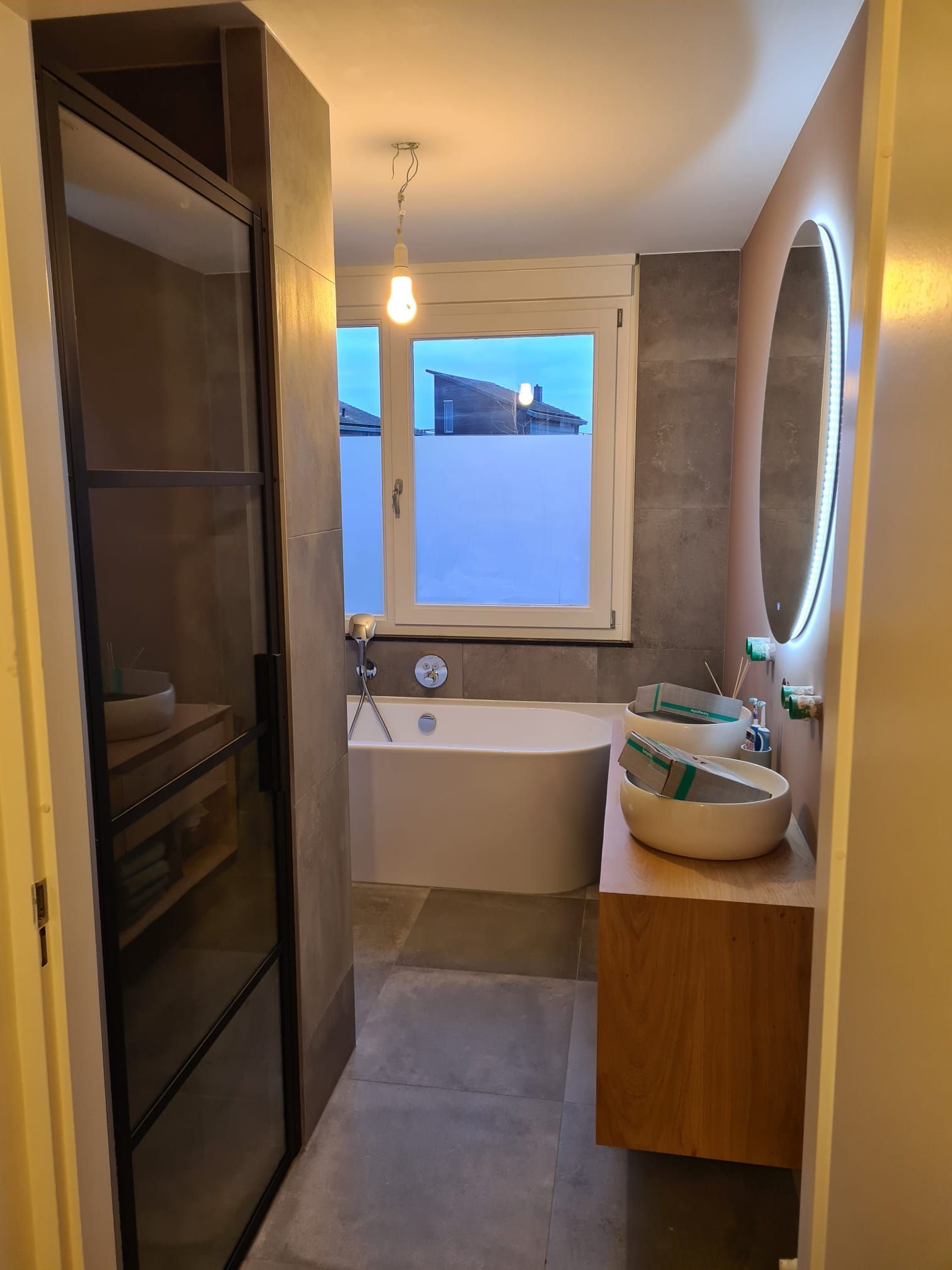 Een moderne badkamer met een wit bad, twee opbouwspoelbakken op een houten badmeubel, een grote ronde spiegel en grijze tegels.