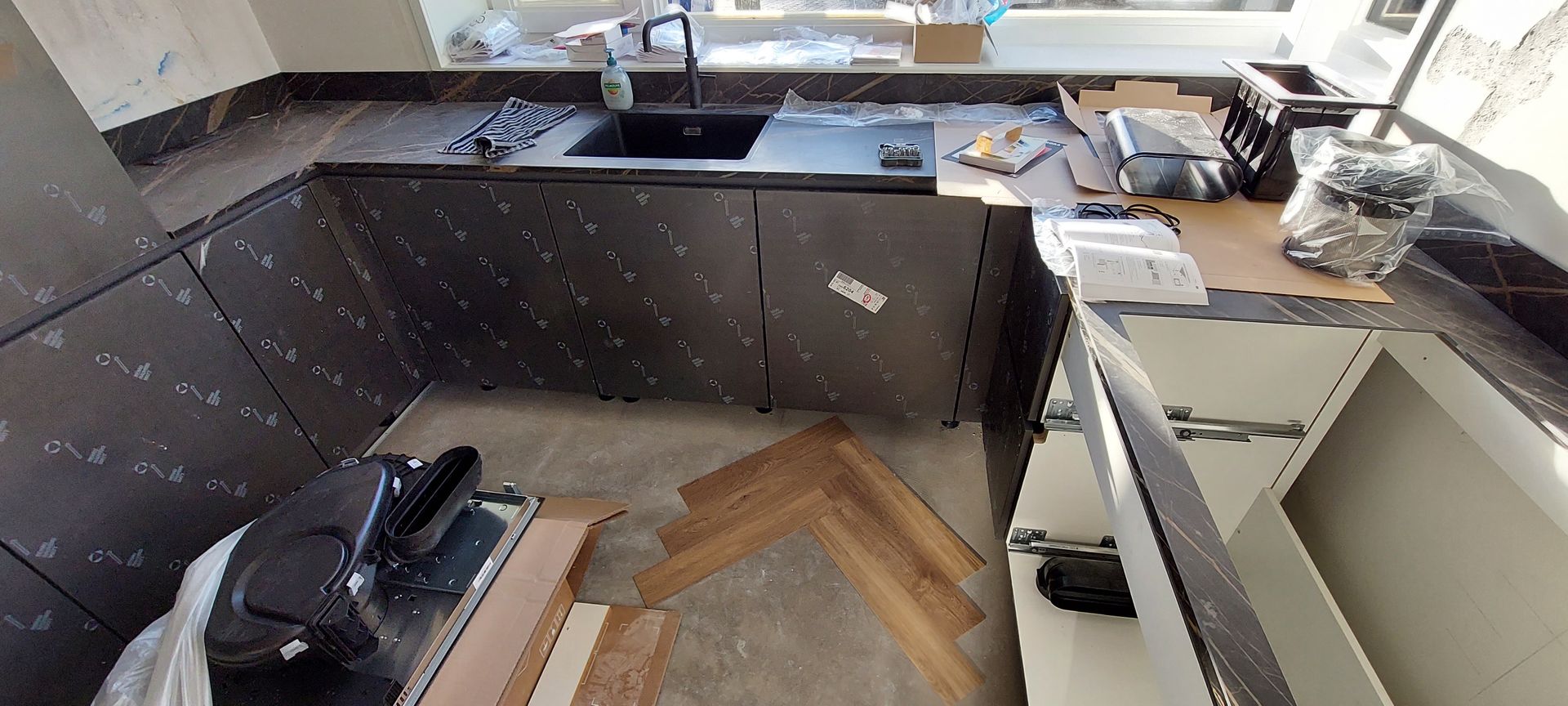 Een keuken in aanbouw met donkere kasten, een zwarte spoelbak en een visgraatparketvloer die nog gelegd moet worden.