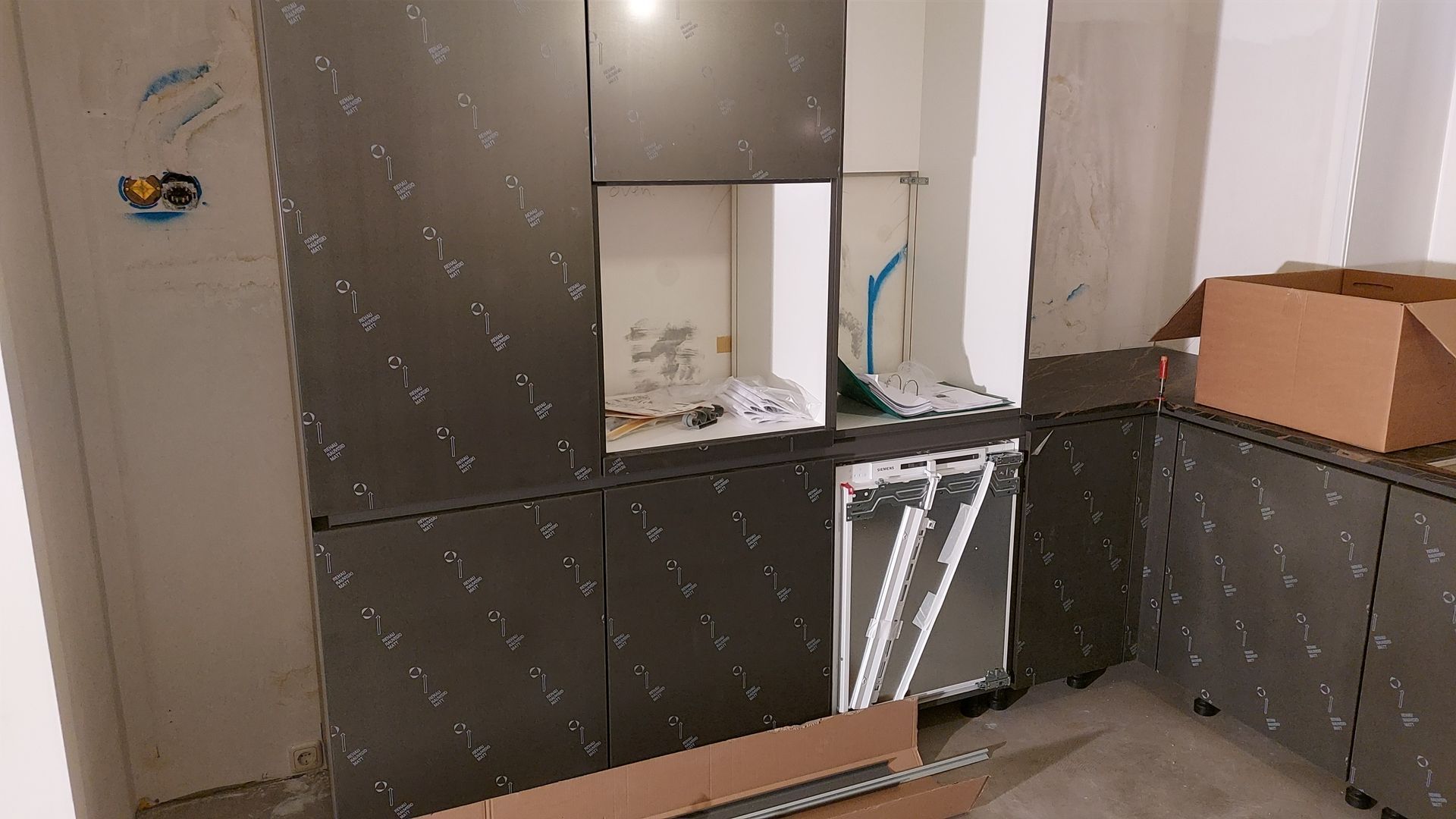Een keuken in aanbouw met donkergrijze kastjes, een ingebouwde vaatwasser en een kartonnen doos op het aanrecht.