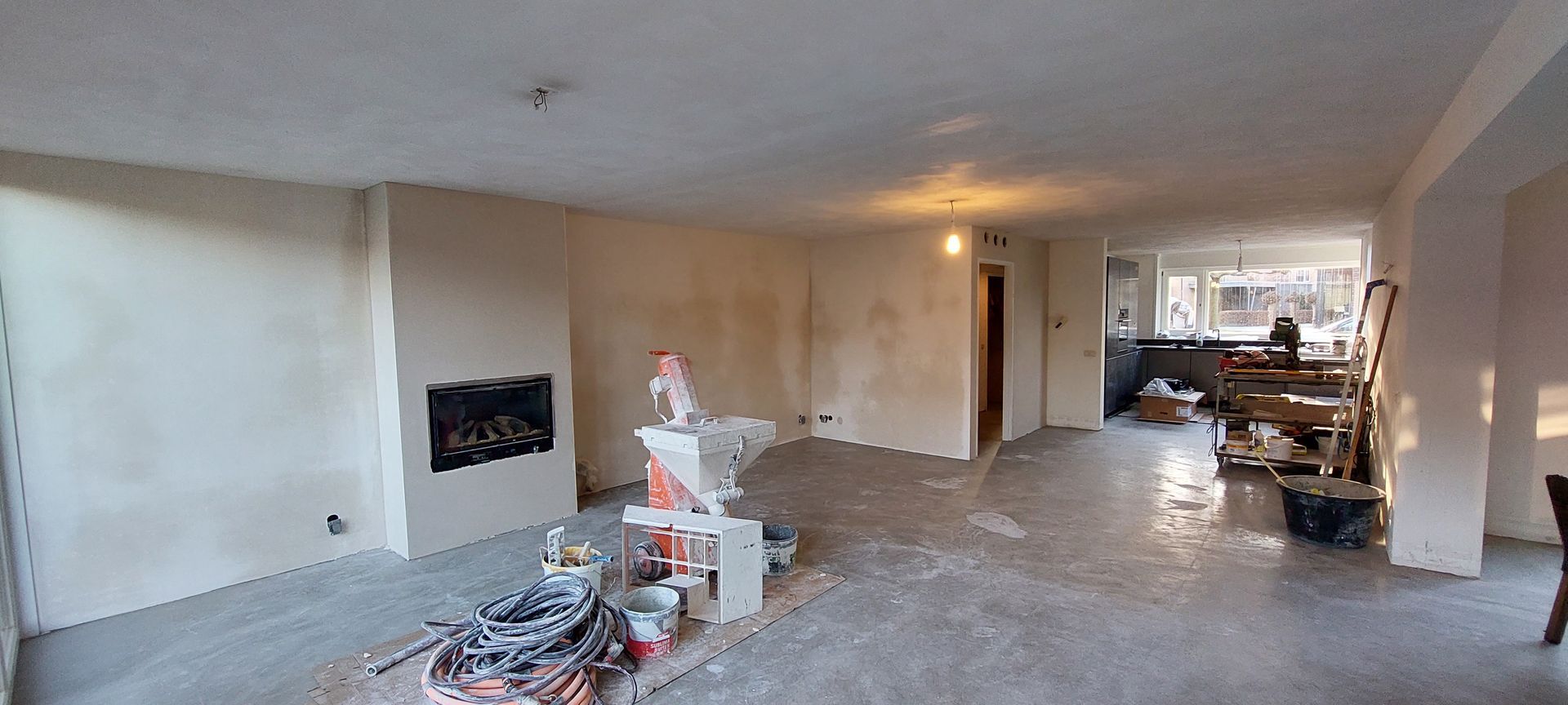 Een interieurfoto van een kamer die wordt gerenoveerd, met nieuw gestucte muren, een centrale open haard en bouwmachines.