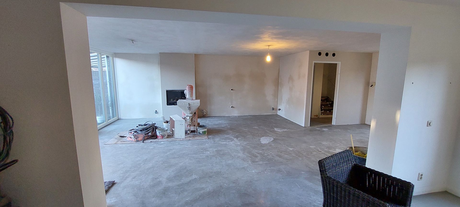 Een overzichtsfoto van een kamer die gerenoveerd wordt, met witte muren, een kale betonnen vloer, een open haard en een deuropening.