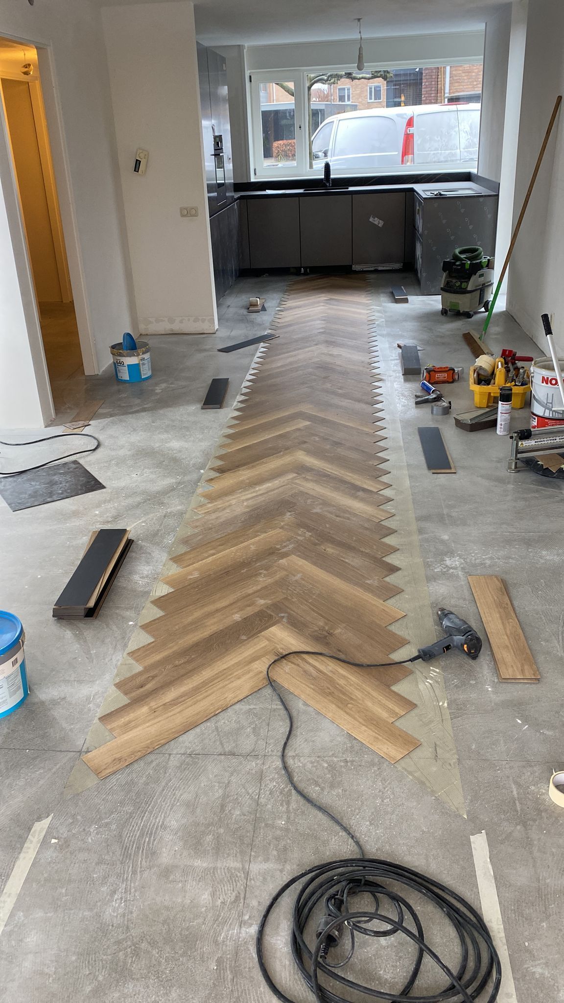 In een keuken die wordt gerenoveerd, wordt een strook houten vloerdelen met visgraatpatroon gelegd op een betonnen ondervloer.
