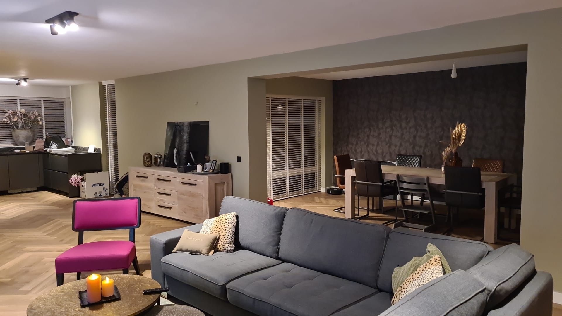 Een moderne woonkamer met een grijze bank, een roze stoel en een eethoek op de achtergrond met een donkere muur met structuur.
