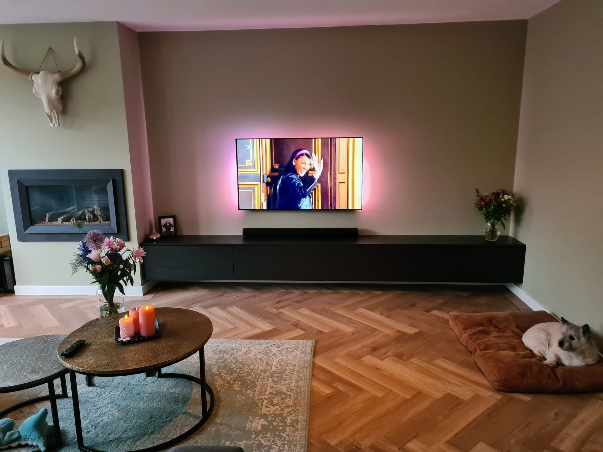Een moderne woonkamer met een aan de muur gemonteerde tv, een lange zwarte console, een open haard, een salontafel en een hond die op een vloerkleed ligt.