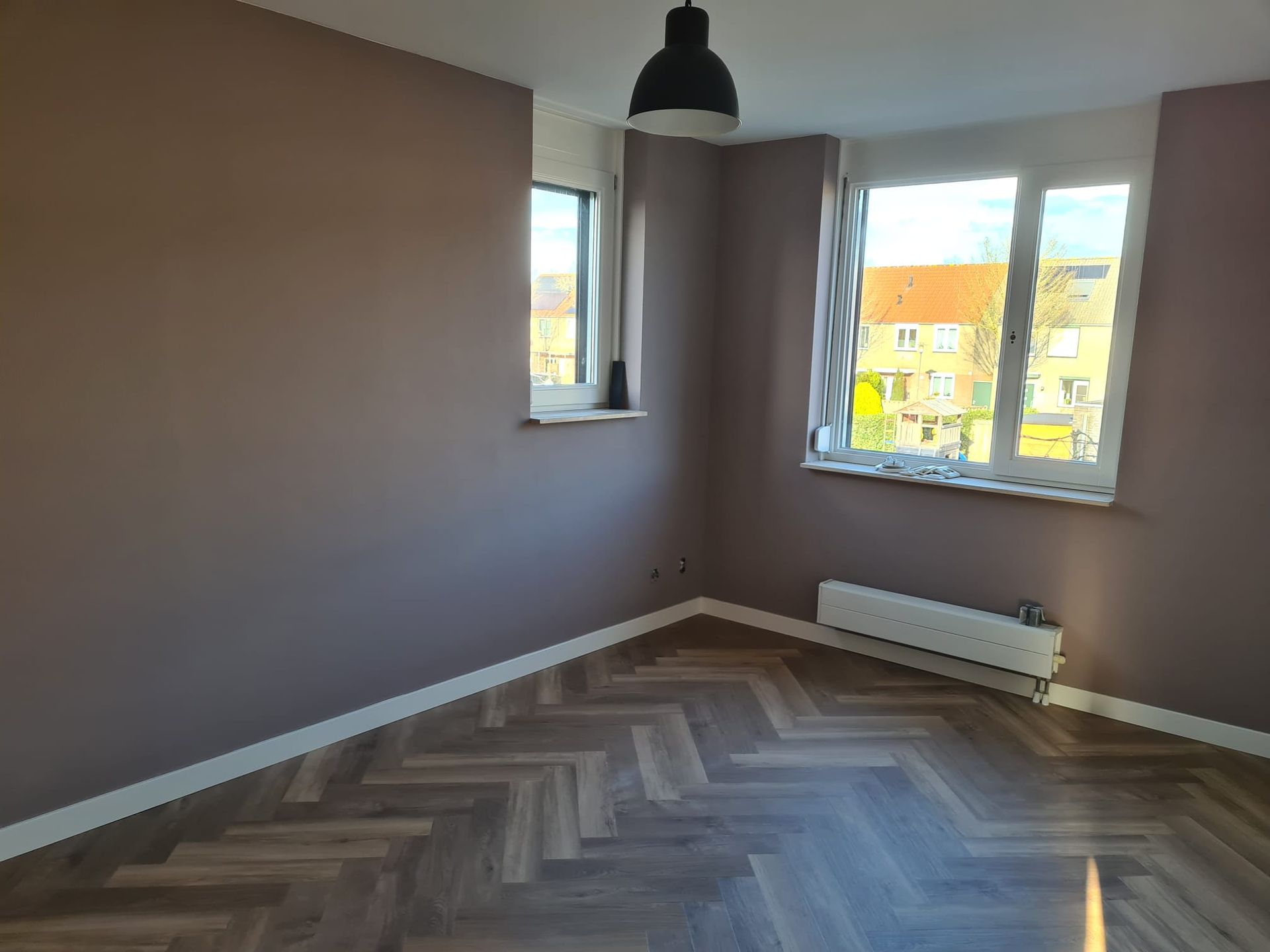 Een lege kamer met stoffig roze muren, een vloer met visgraatpatroon, een groot raam, een radiator en een hanglamp.