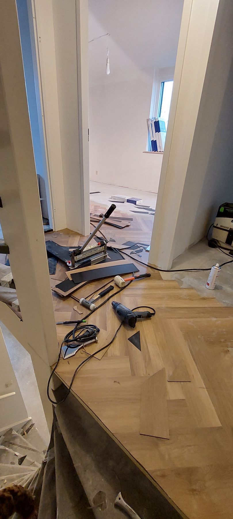 Werk in uitvoering: een kamer met onafgewerkte houten vloer in visgraatpatroon, verspreid gereedschap en een deuropening naar een andere ruimte.