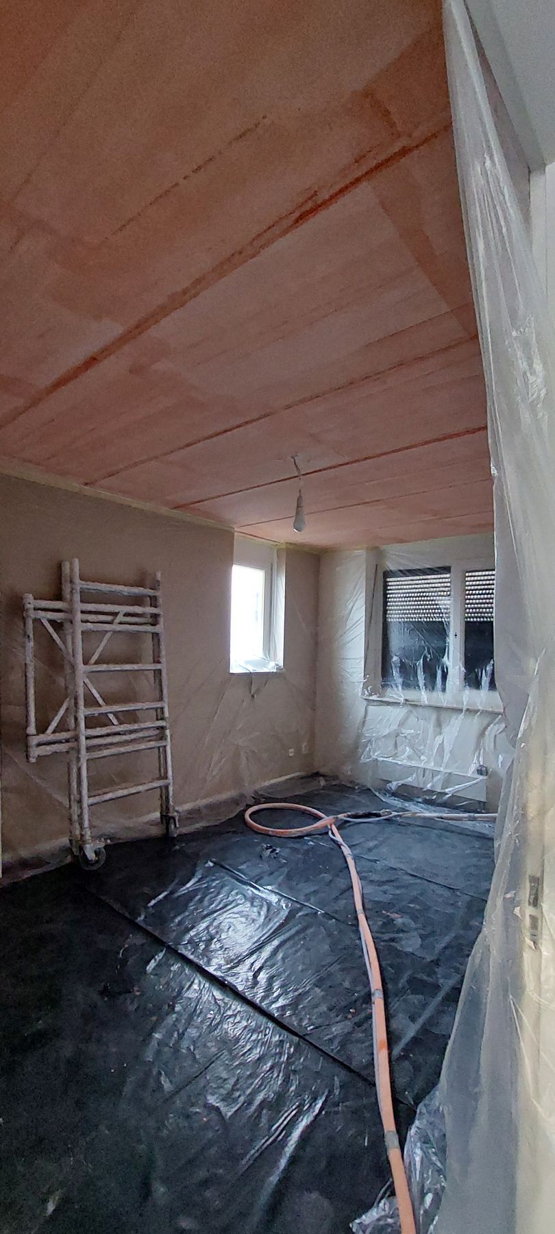 Een onafgewerkte kamer in renovatie met een houten plafond, gestucte muren, steigers en een zwarte plastic vloerbedekking.