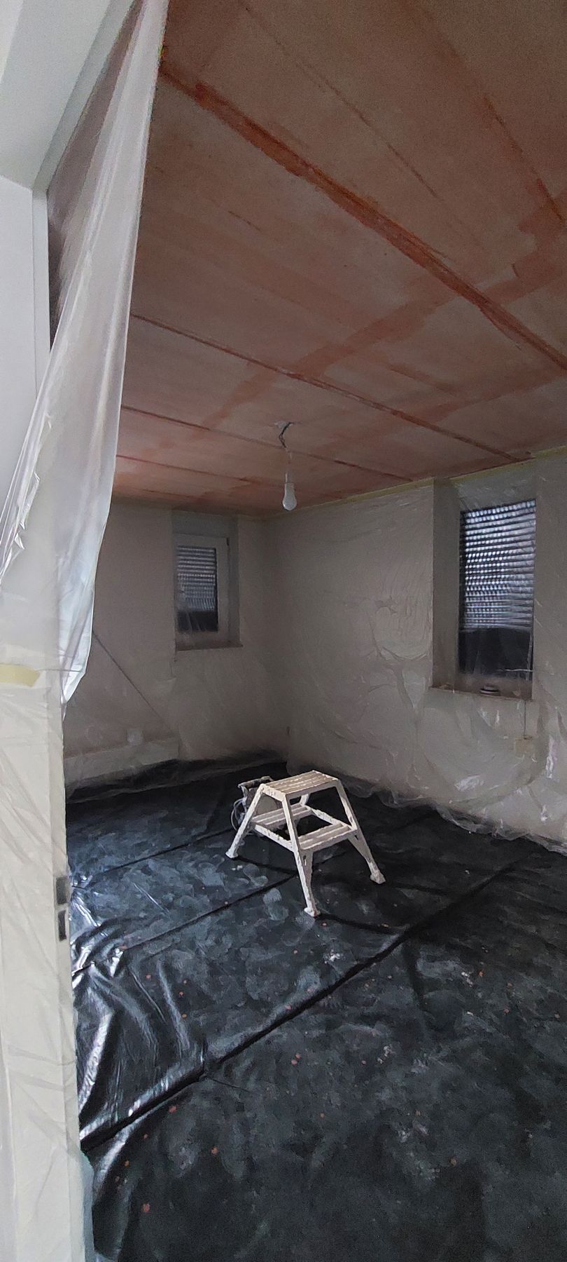 Een onafgewerkte kamer met een plafond van multiplex, lichtgekleurde muren, ramen en een beschermend zwart zeil over de vloer.