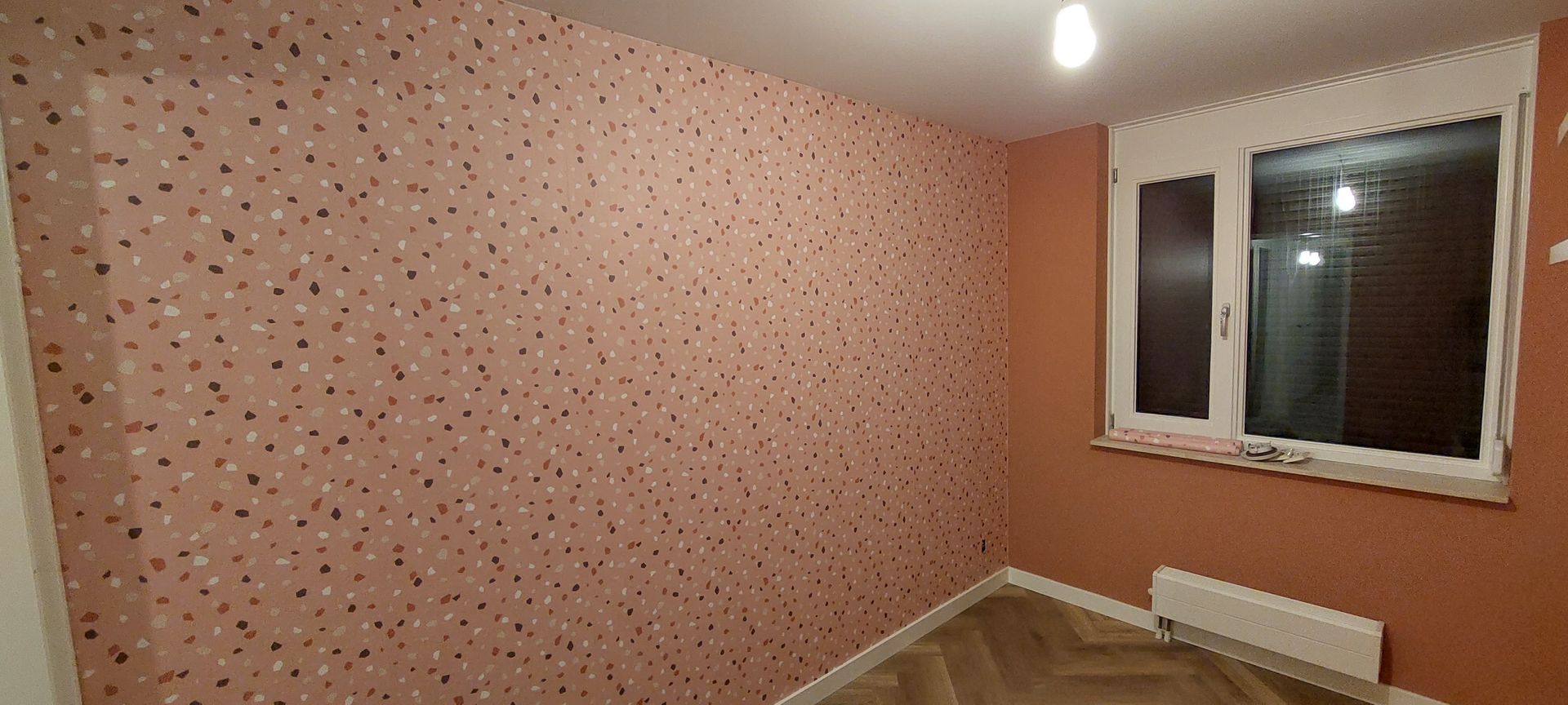 Een kamer met één muur bekleed met roze behang met een confetti-patroon en een aangrenzende terracotta-kleurige muur met een raam.