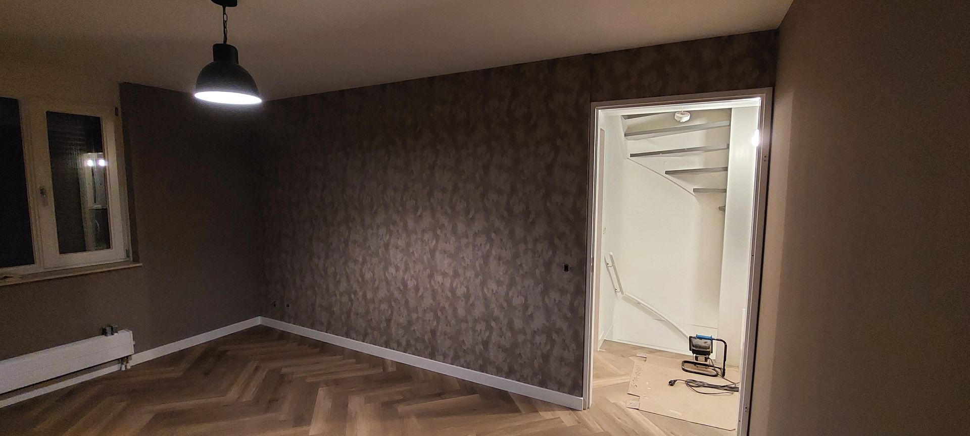 Een binnenkamer met donker behang met patroon, een houten vloer in visgraatpatroon, een enkele hanglamp en een open deuropening.