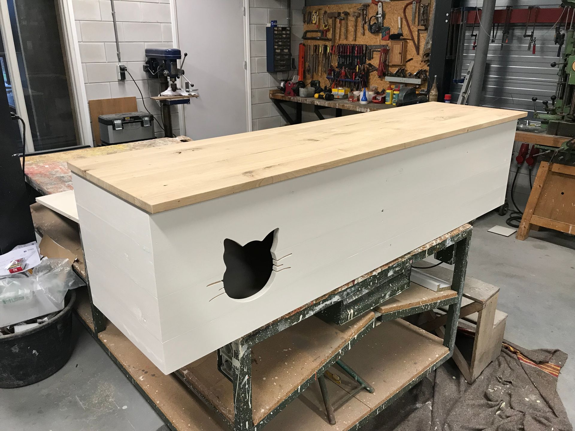 Een lange, witte, rechthoekige houten doos met een uitsparing in de vorm van een kat, die op een werkbank in een houtwerkplaats staat.