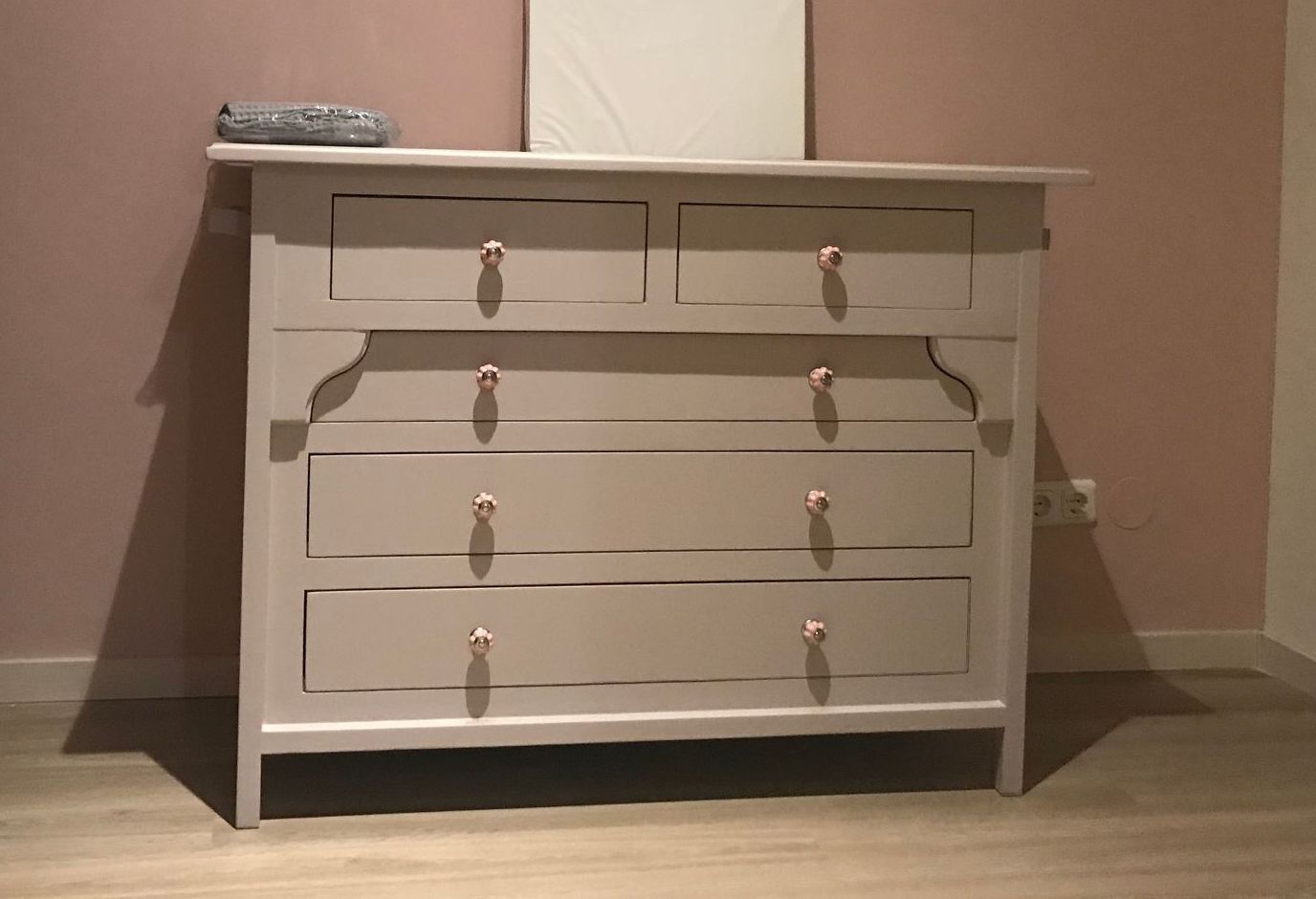 Een lichtroze houten dressoir met vier lades en kleine, ronde witte knopjes, tegen een bijpassende roze muur.