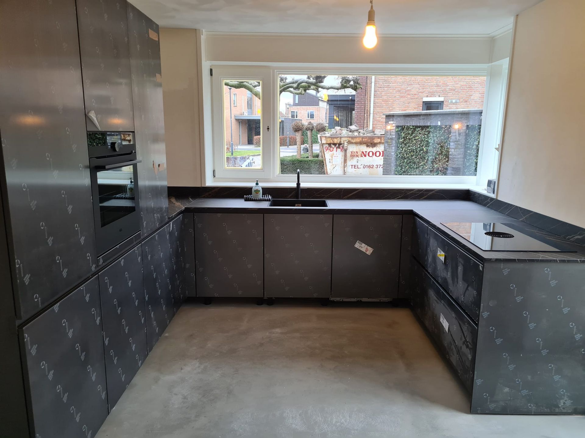 Een U-vormige keuken met donkere, gedecoreerde kasten, een donker aanrechtblad, een spoelbak en een ingebouwde oven onder een groot raam.