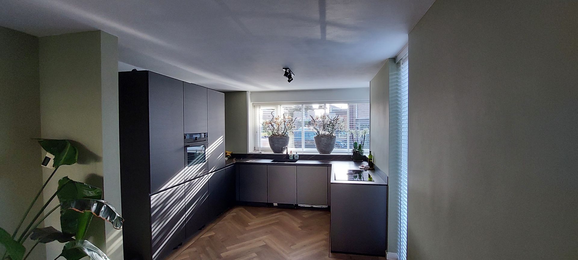 Een moderne keuken met zwarte kasten, een visgraatparketvloer en een groot raam met twee potplanten.