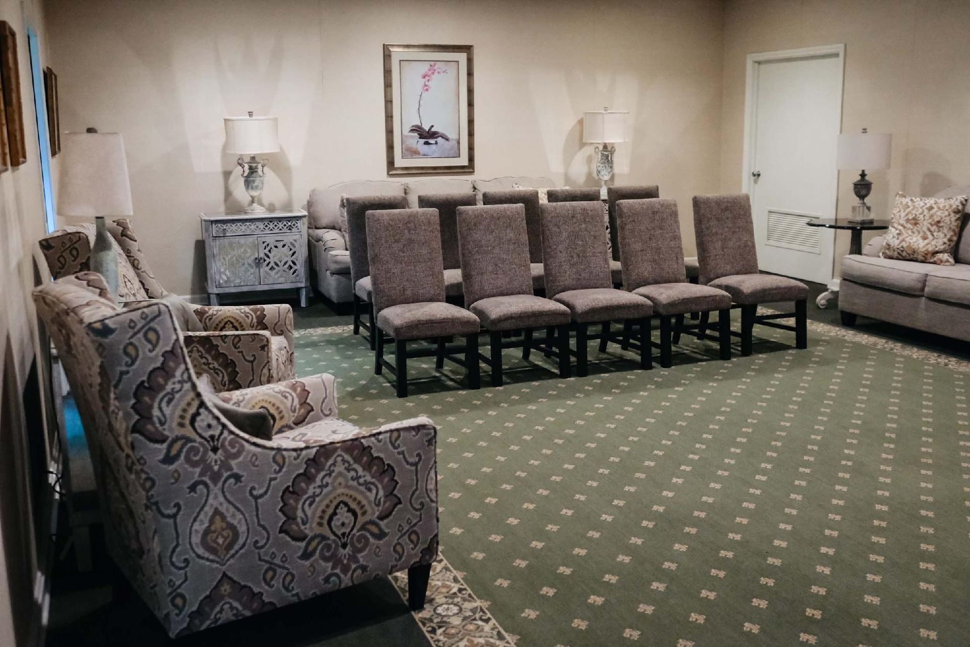 An empty room with chairs arranged for a service, featuring neutral tones, lamps, and a patterned rug.