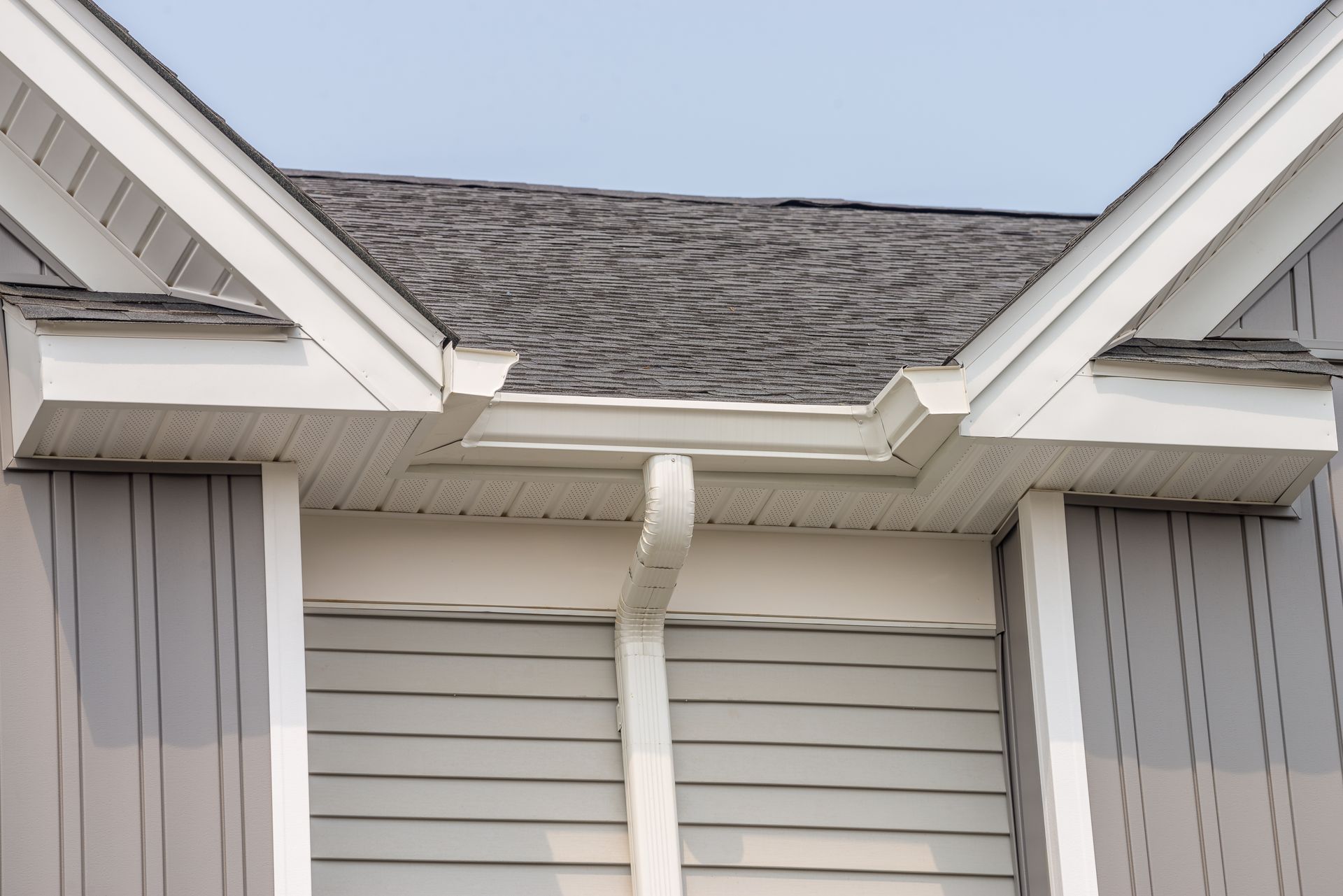 Roof eaves with a white gutter and downspout above gray siding and windows