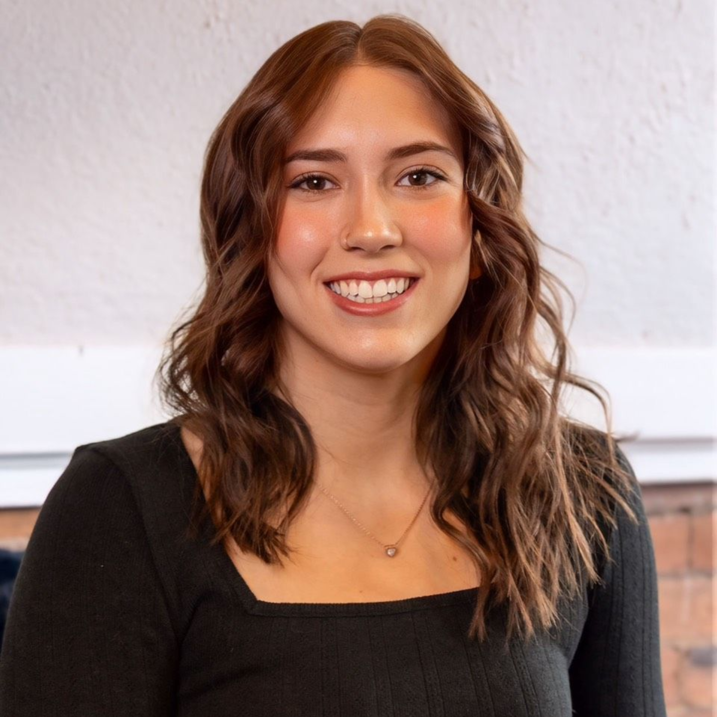 Robin McIntyre — Laramie, Wy — Luna Salon & Spa Smiling person with wavy brown hair in a black top against a light wall