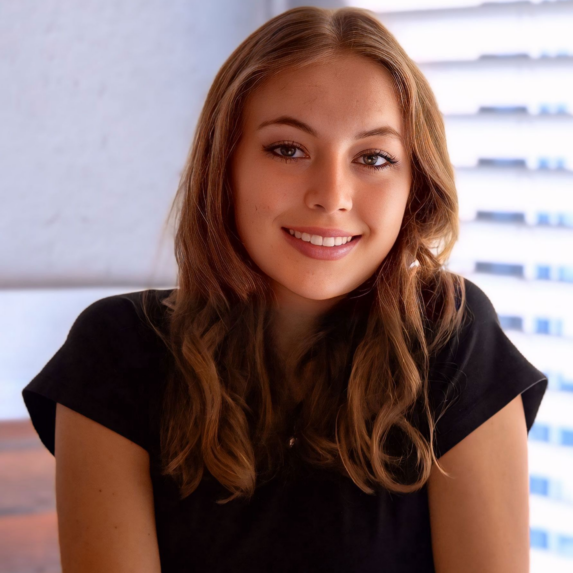 Chayse Graham — Laramie, Wy — Luna Salon & Spa Smiling woman in a black shirt sitting indoors near a bright window with blinds