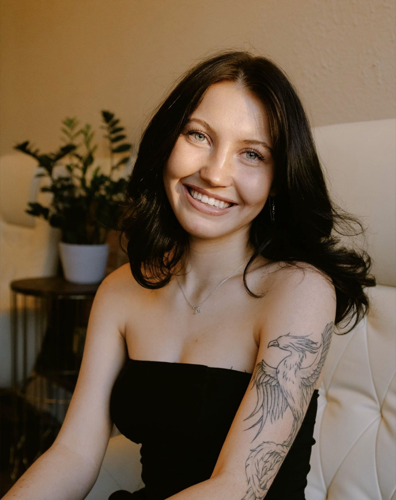 Kierra Osborne — Laramie, Wy — Luna Salon & Spa Smiling woman with wavy brown hair in a black top, seated against a brick and white wall.