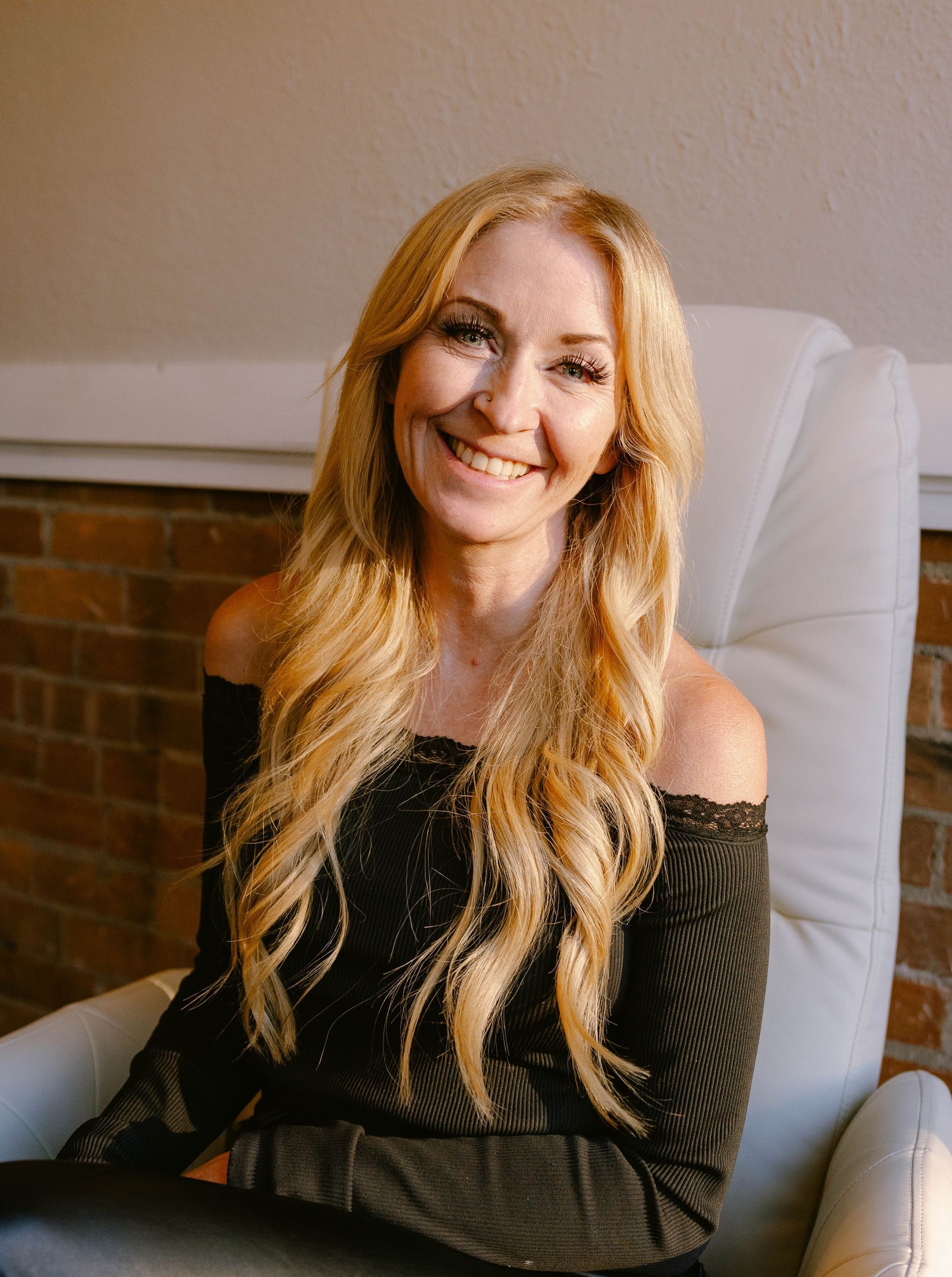 Jen Vincent — Laramie, Wy — Luna Salon & Spa Smiling blonde woman in a black shirt indoors, with soft window light and white blinds behind her