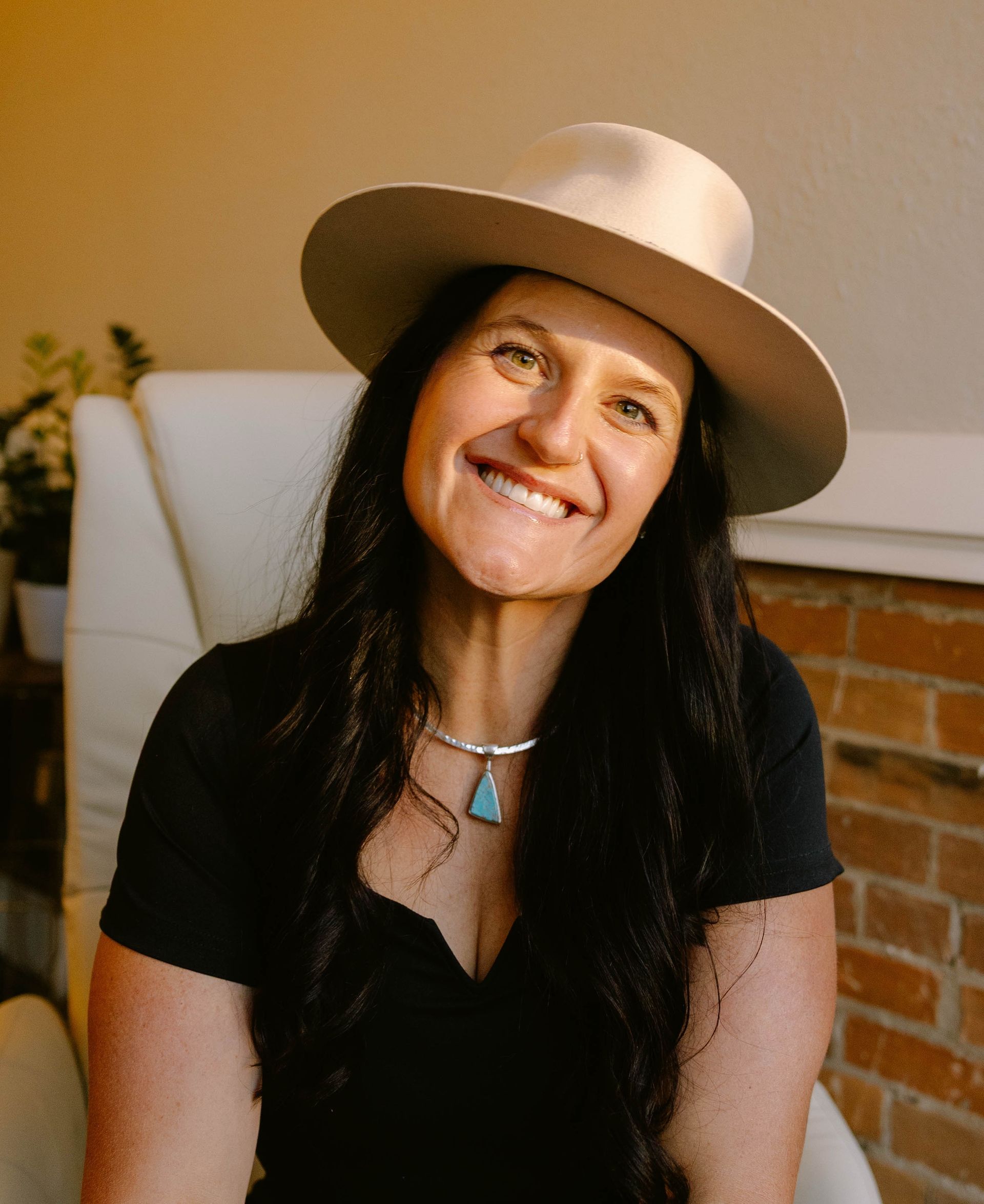 Chelsey Moon — Laramie, Wy — Luna Salon & Spa Smiling woman in a red satin blouse seated indoors against a brick and white wall.