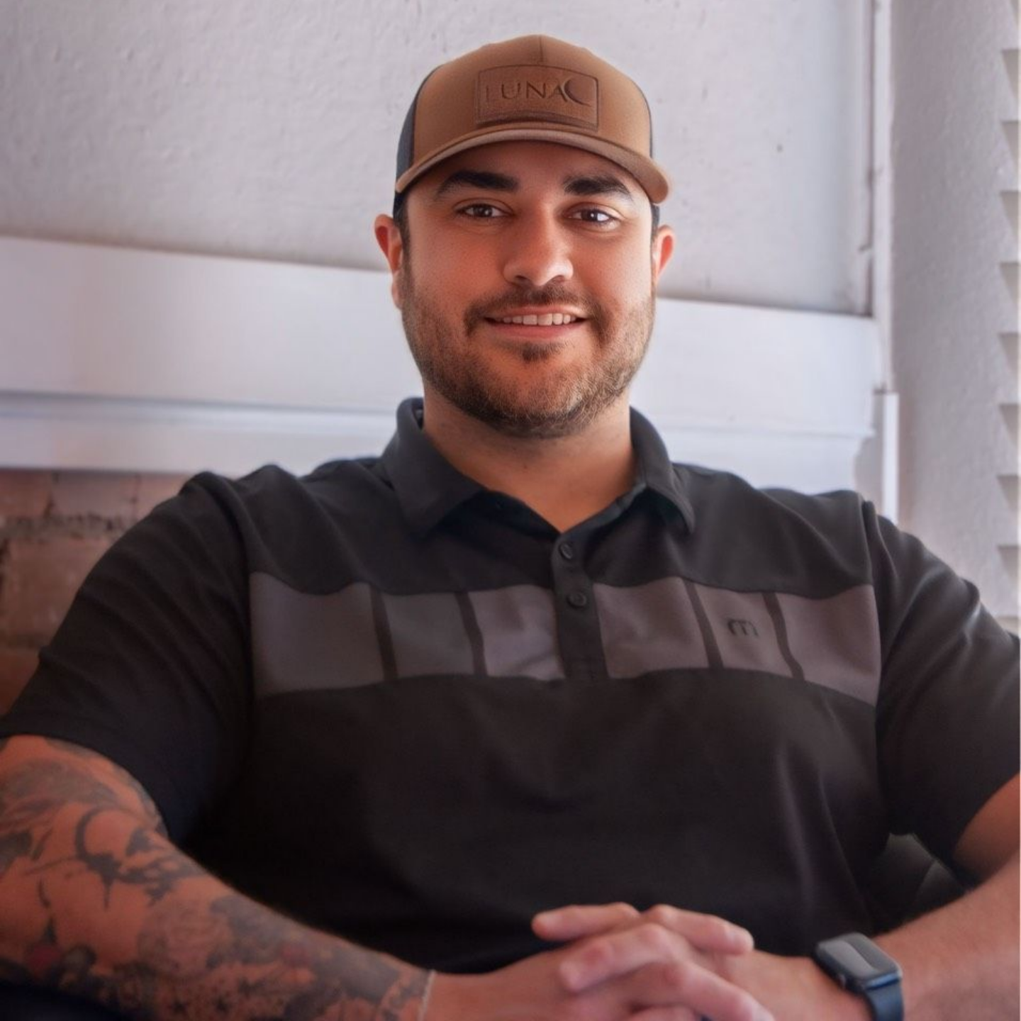 Fernando Zamora — Laramie, Wy — Luna Salon & Spa Man seated indoors, smiling with folded hands, wearing a brown cap and black polo shirt.