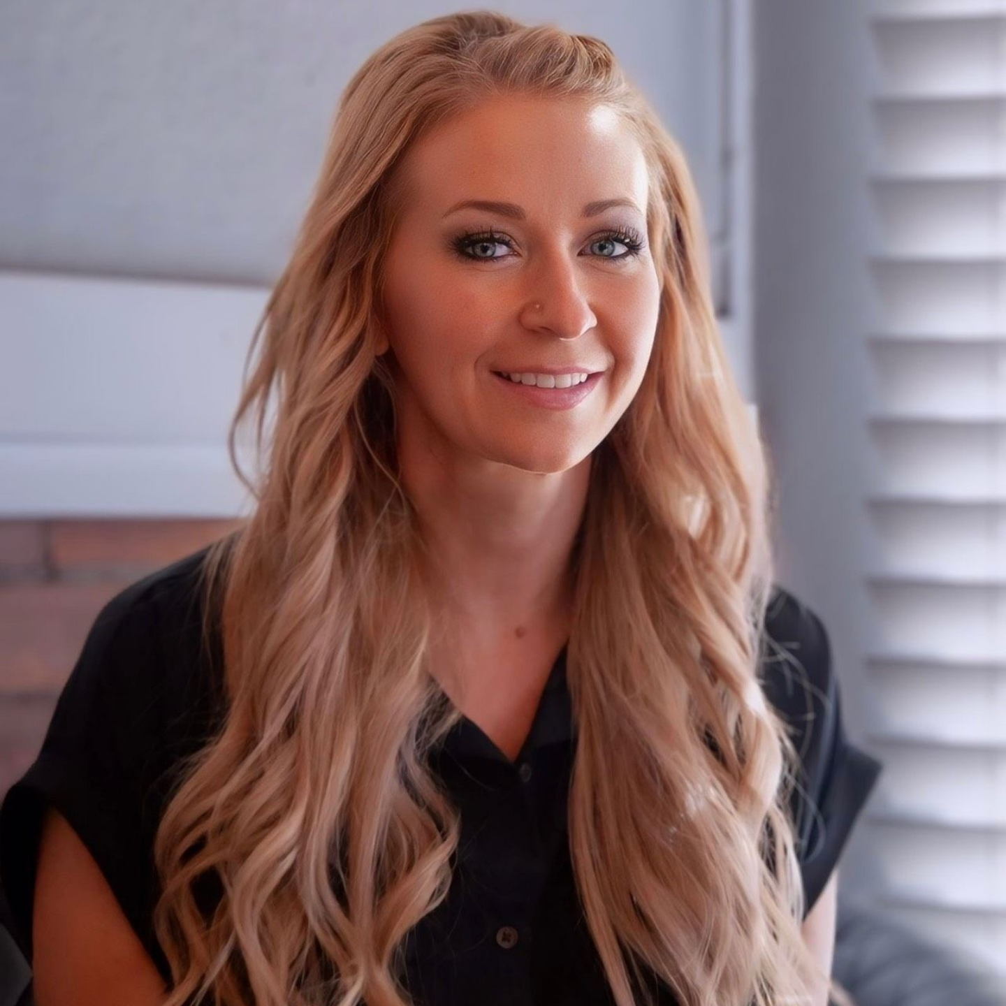 Jen Vincent — Laramie, Wy — Luna Salon & Spa Smiling blonde woman in a black shirt indoors, with soft window light and white blinds behind her