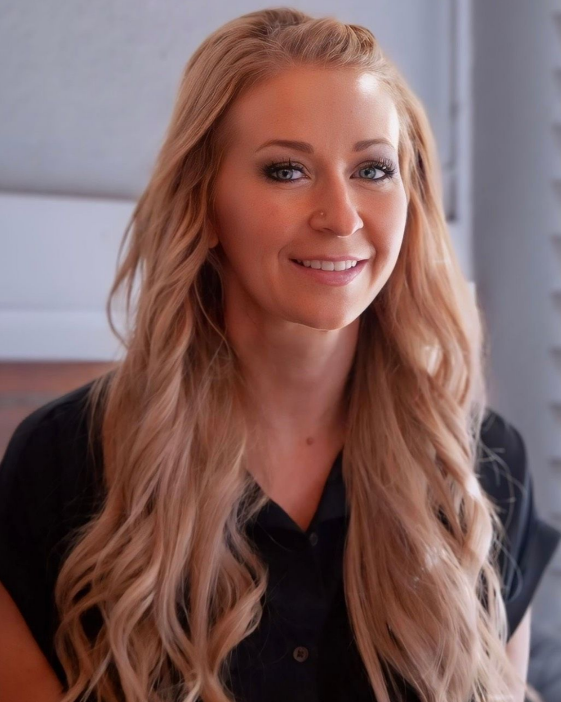 Smiling woman with long wavy blonde hair in a black blouse, standing indoors against a light background