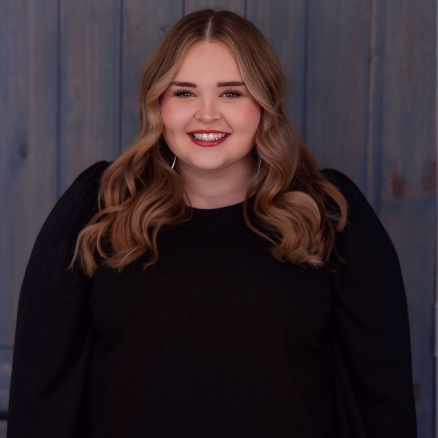 Stormi Love — Laramie, Wy — Luna Salon & Med Spa Smiling person with wavy hair wearing a black top against a blue-gray wooden backdrop