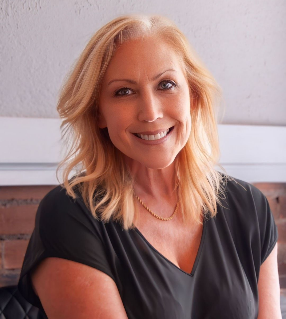 Smiling woman with blonde hair in a black top, seated against a light wall.