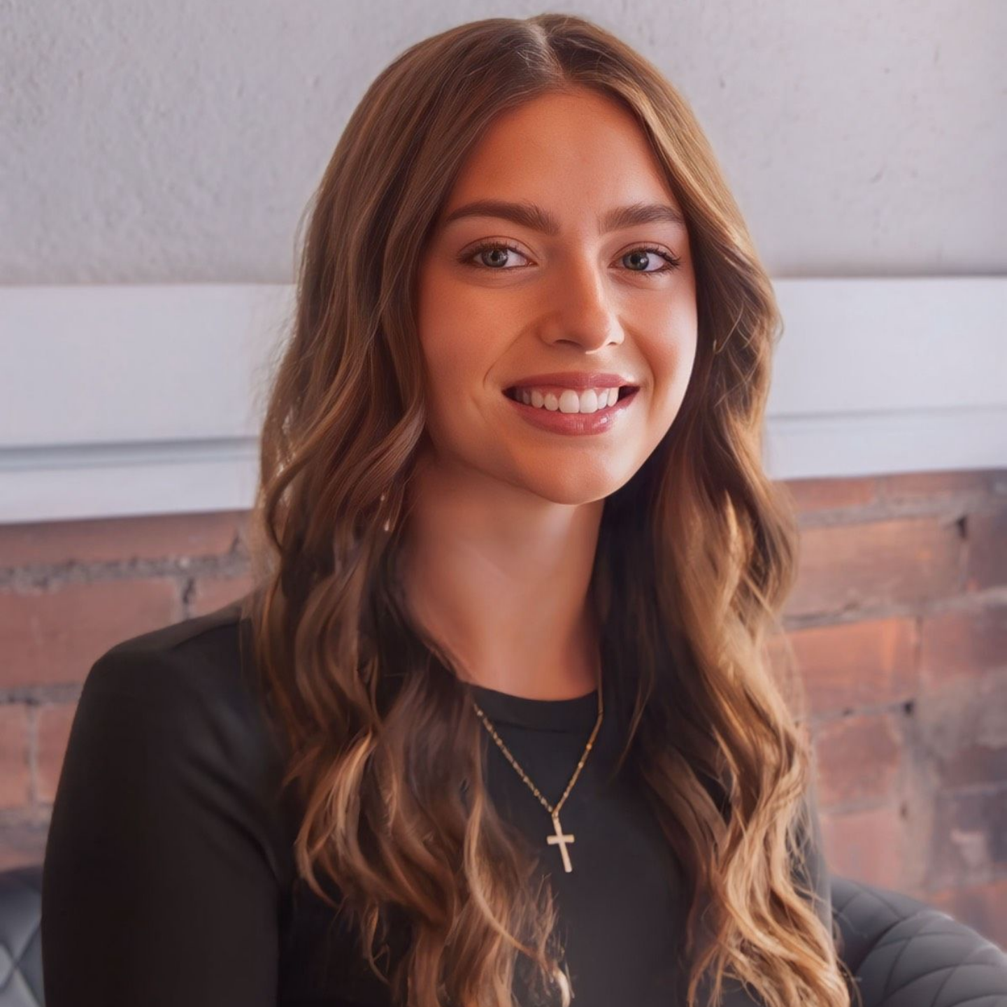 Connor Kathleen — Laramie, Wy — Luna Salon & Spa Smiling woman with long wavy hair, wearing a black top and cross necklace, seated indoors against a brick wall.