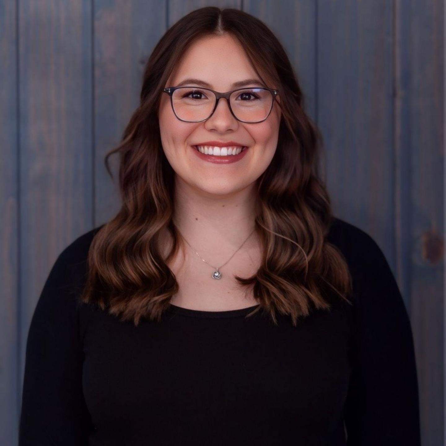 Picture of a Girl with a Black Shirt and Blonde Hair — Laramie, Wy — Luna Salon & Med Spa Portrait of a smiling person with glasses and wavy brown hair against a blue-gray wooden backdrop