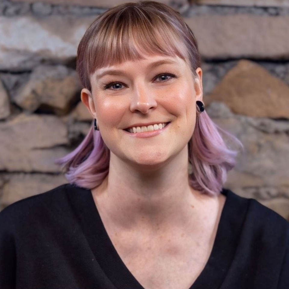Coming Soon Smiling person with lavender hair against a stone wall background