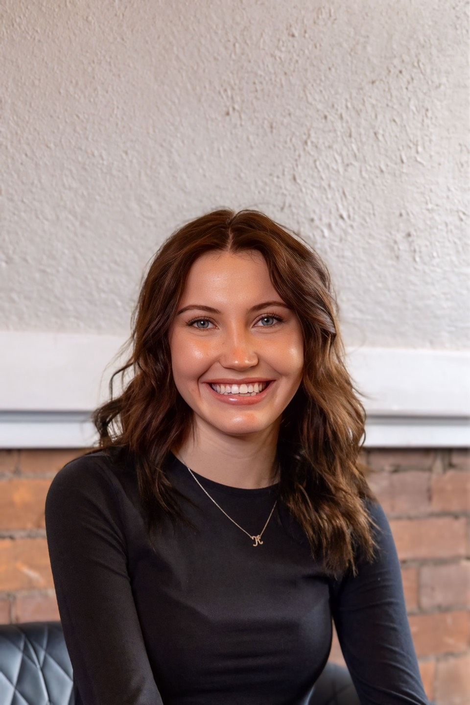 Smiling woman in a black top seated indoors against a white wall and brick backdrop