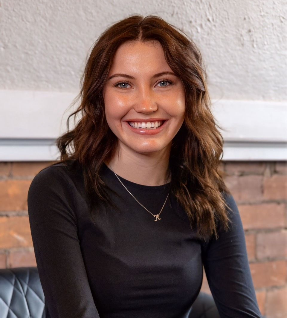 Kierra Osborne — Laramie, Wy — Luna Salon & Spa Smiling woman with wavy brown hair in a black top, seated against a brick and white wall.
