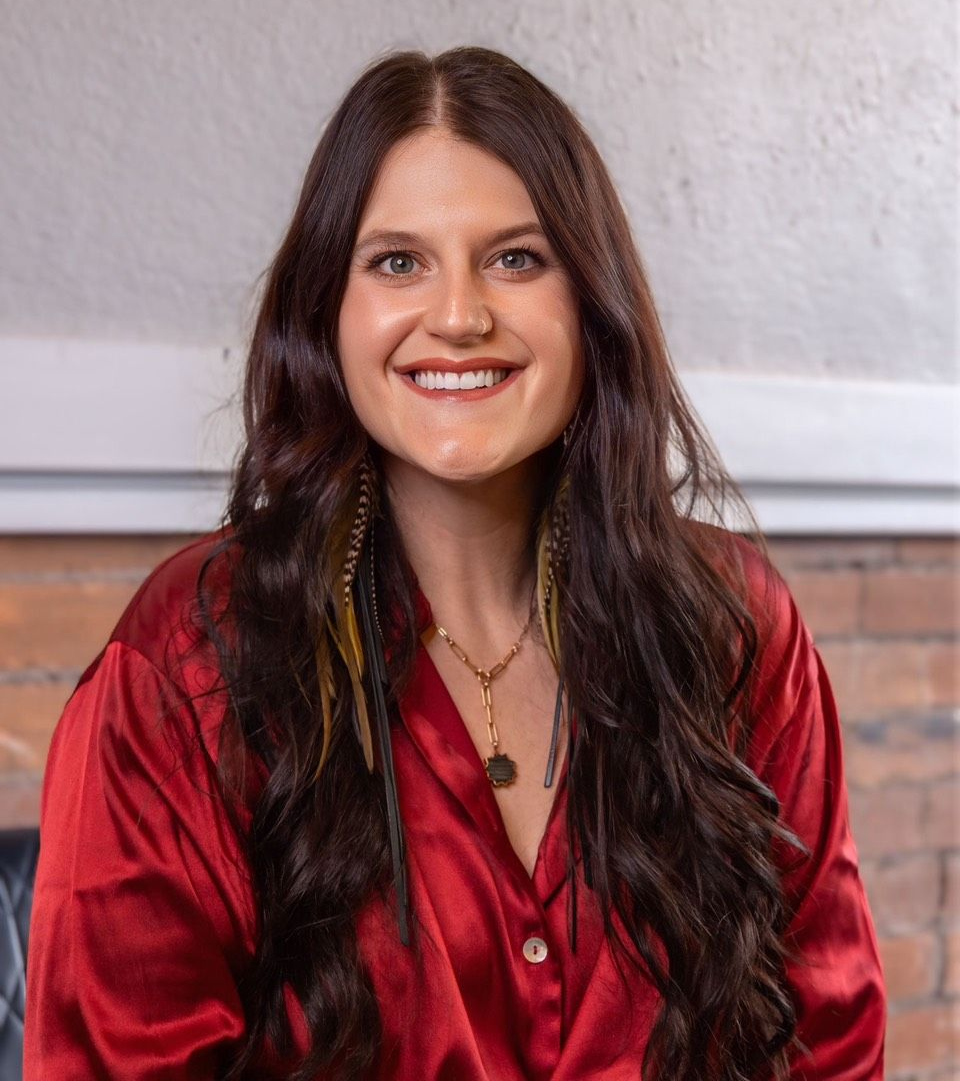 Chelsey Moon — Laramie, Wy — Luna Salon & Spa Smiling woman in a red satin blouse seated indoors against a brick and white wall.