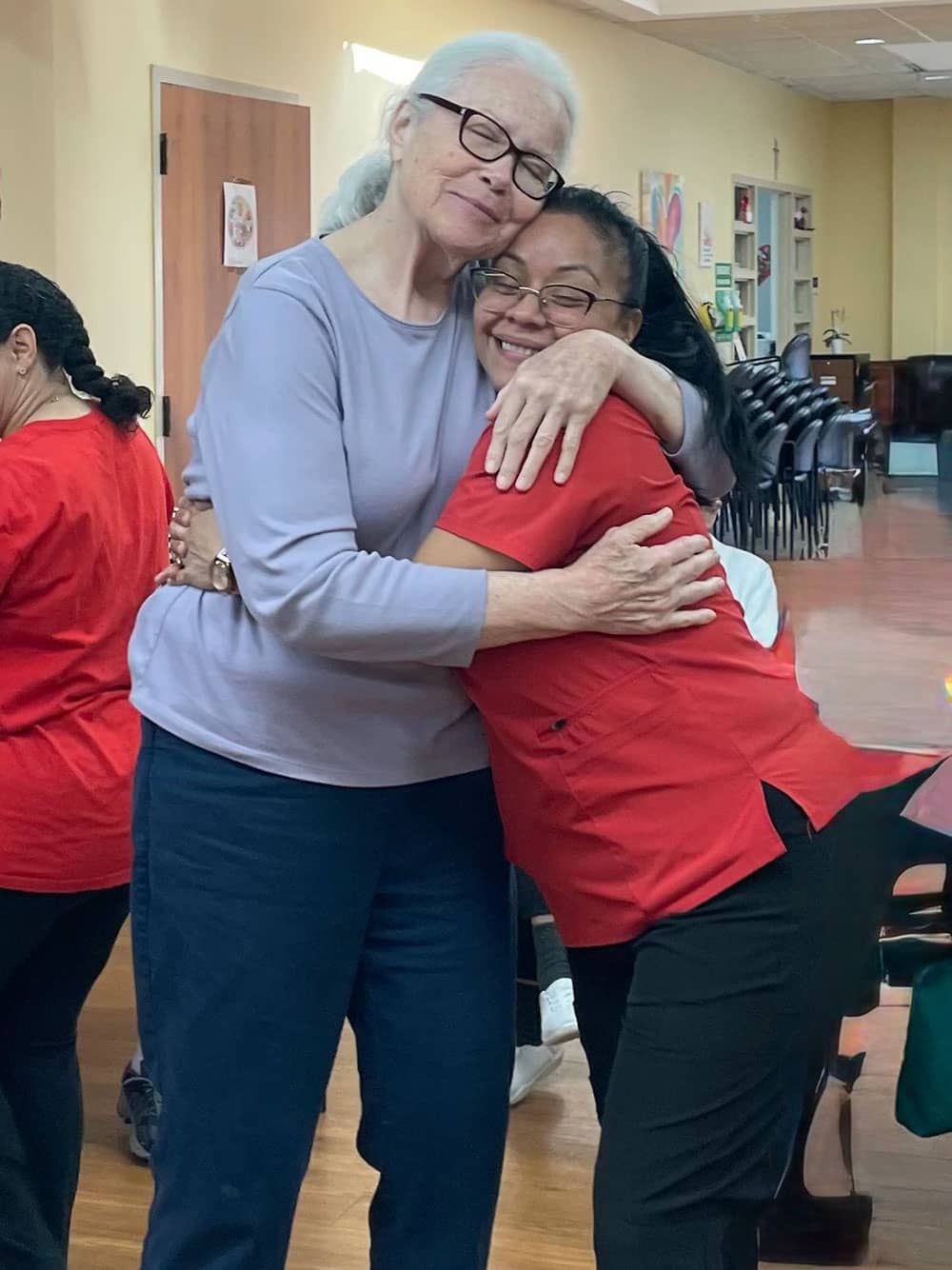 Two people hugging in a hallway, one in a red shirt and one in a lavender top, both smiling