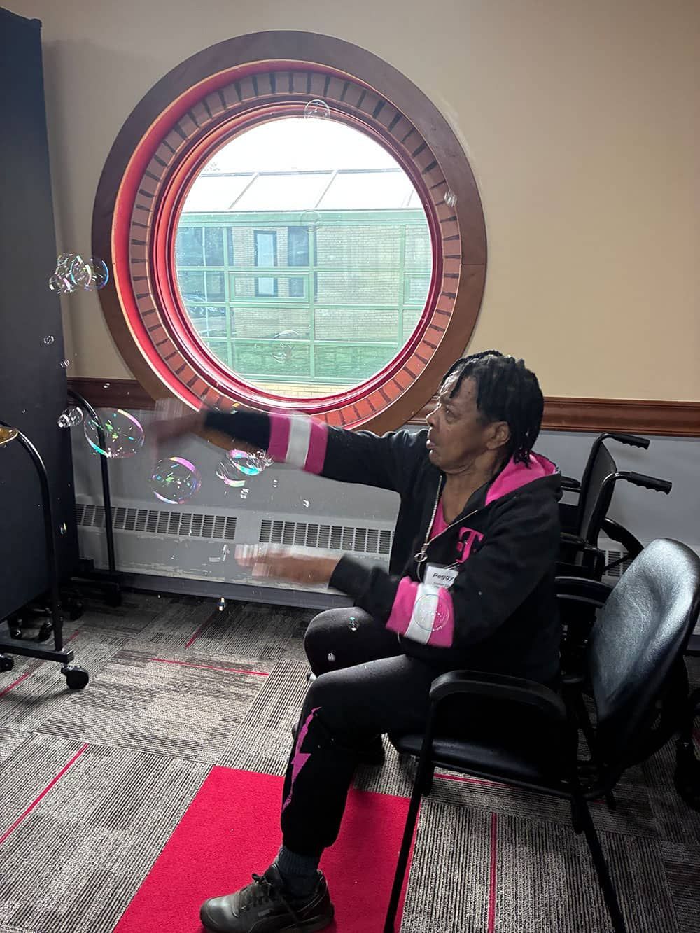 Person seated indoors on a red rug, reaching toward bubbles near a round window.