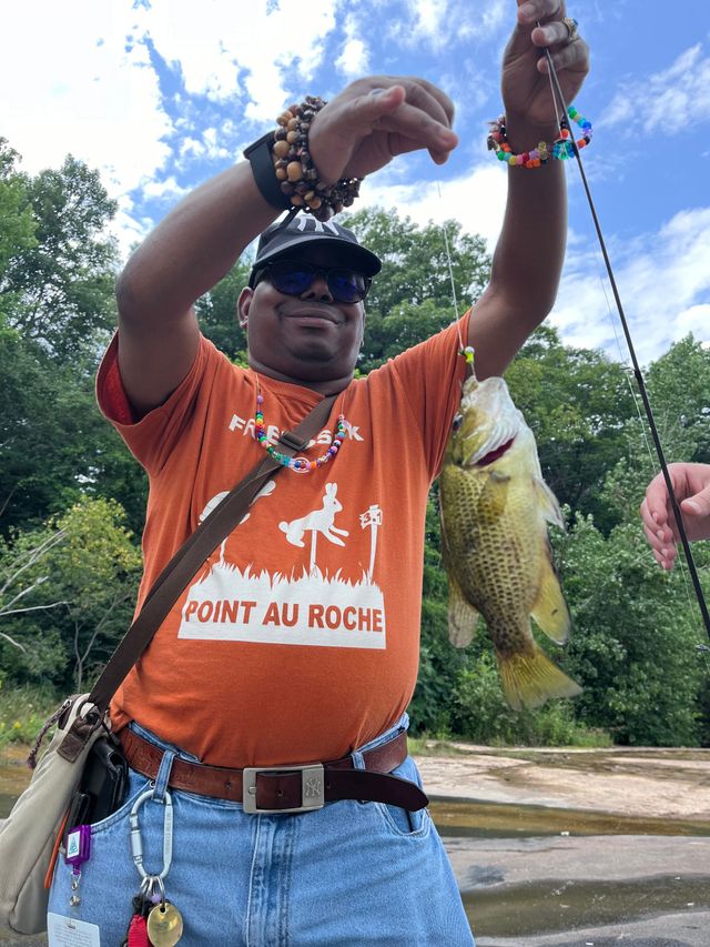 A man in an orange shirt is holding a fish on a fishing pole he caught.