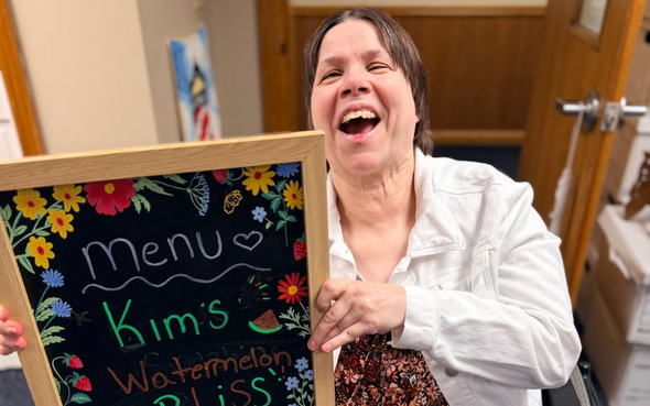 Woman in a wheelchair smiling beside a chalkboard menu in a hallway.