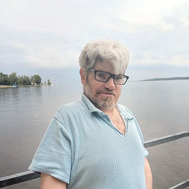 A man with glasses and a beard is standing next to a body of water.
