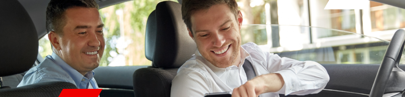 Dois homens sorrindo dentro de um carro. Um está dirigindo, olhando para o celular. O outro está olhando para a frente.