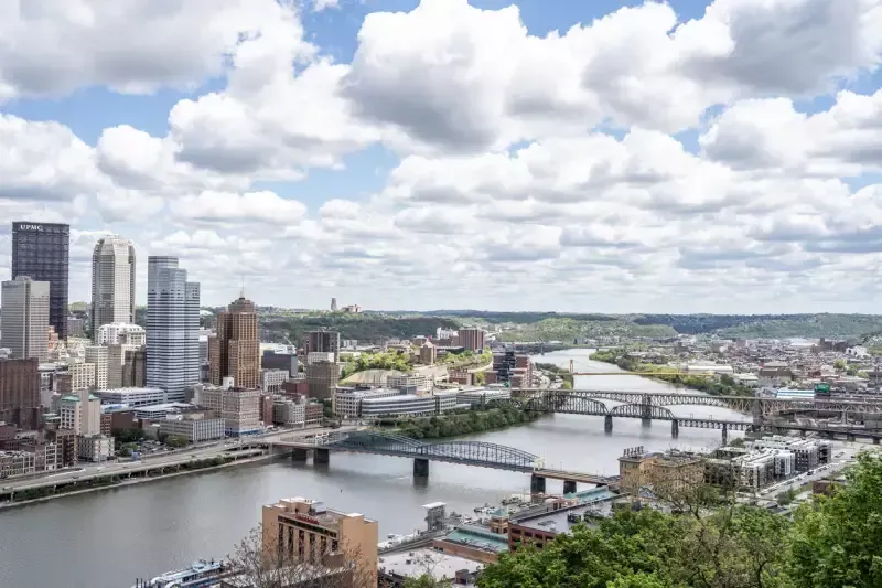 City skyline with rivers and bridges under a partly cloudy sky. Tall buildings and trees line the riverbanks.