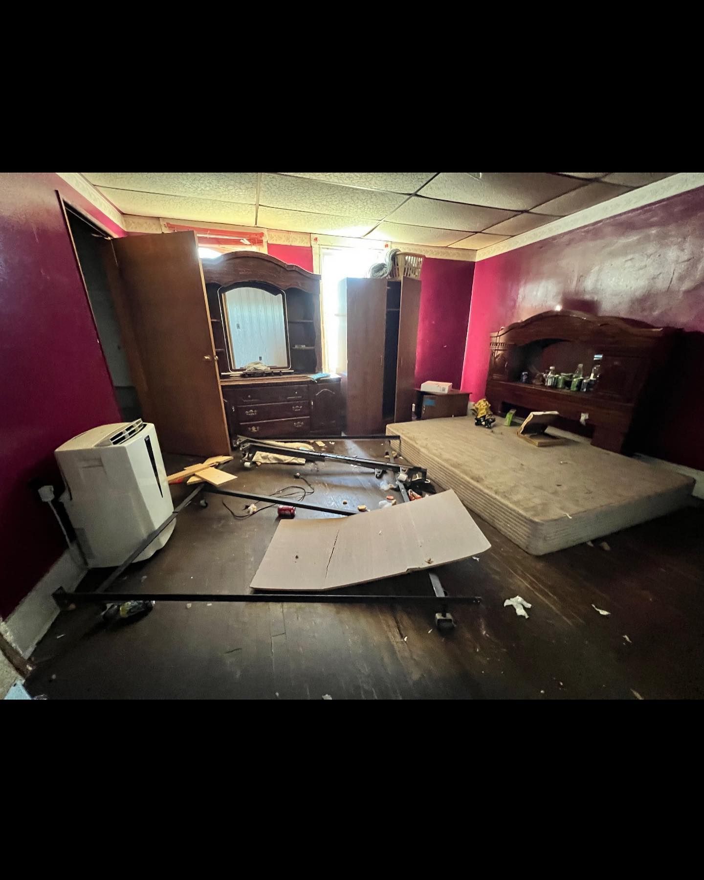 A cluttered, damaged bedroom with a bed, dresser, and debris scattered on the floor; pink walls and a damaged ceiling.