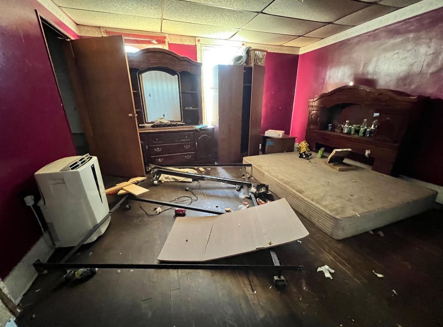 A cluttered bedroom with damaged furniture, debris on the floor, and pink walls. A bed, dresser, and cabinet are visible.
