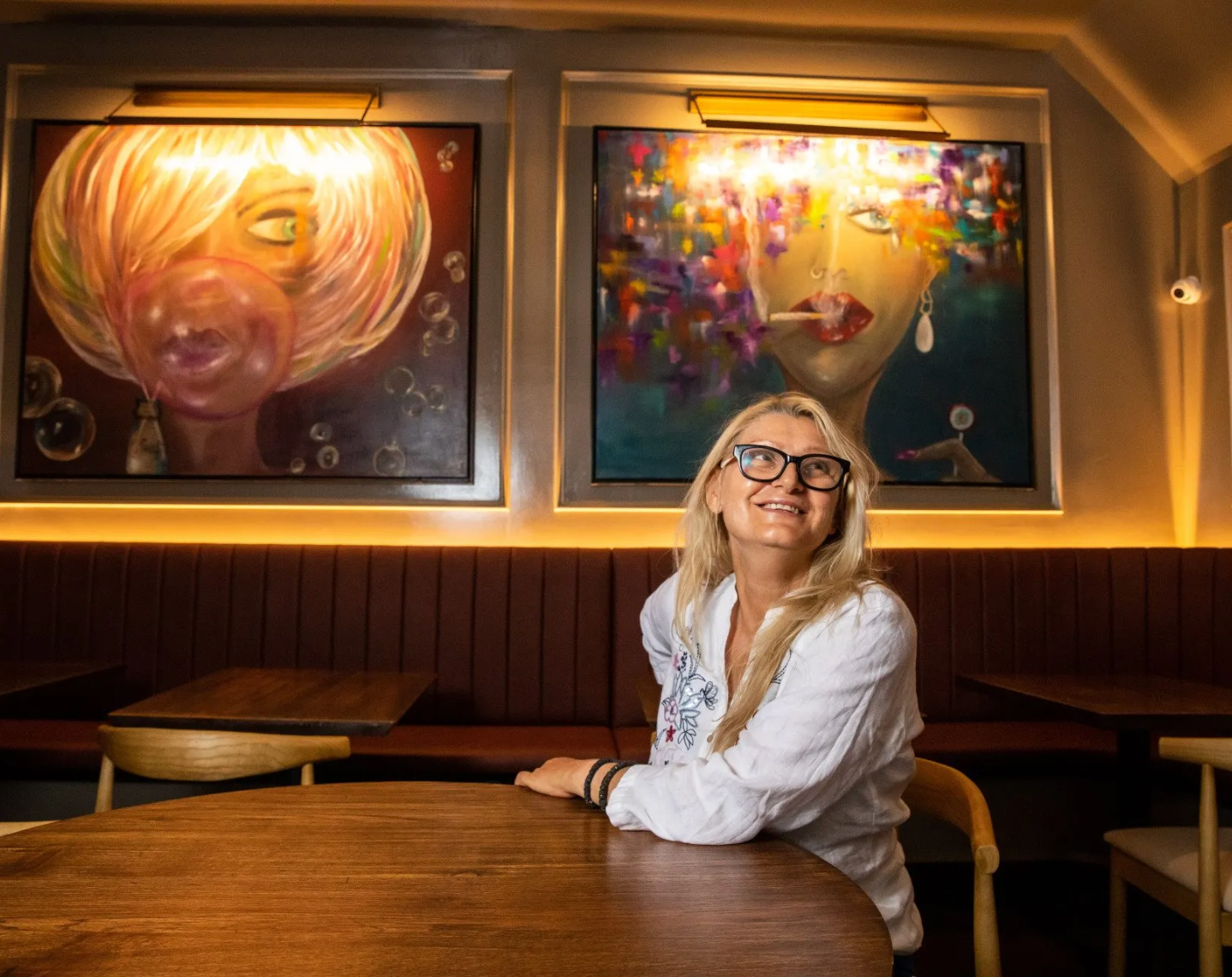 Svetlana Alimpijevic smiles at a table in a restaurant, beneath two abstract paintings.