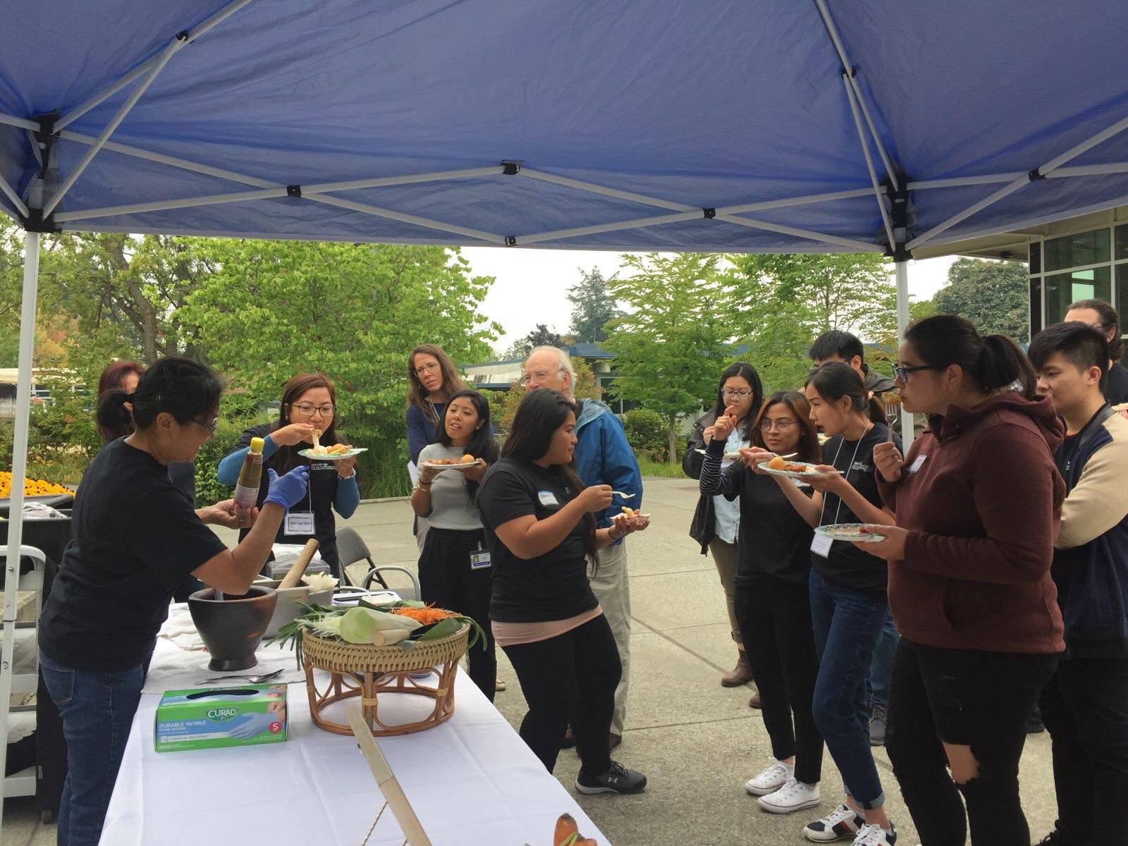 A group of people are standing around a table eating food under a blue tent.